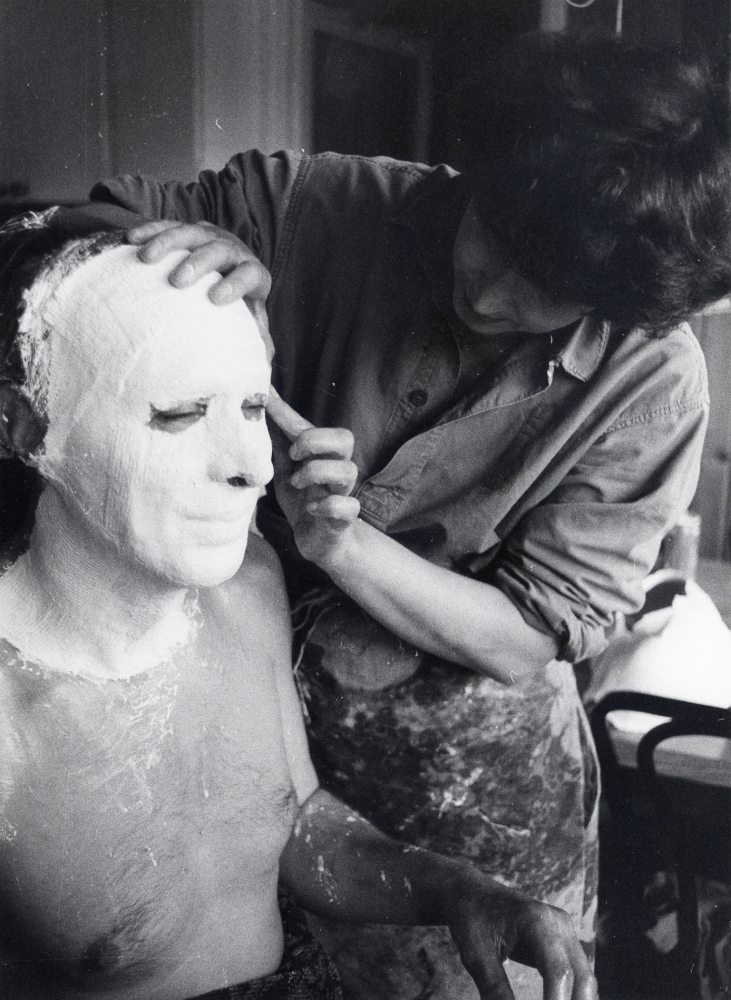 Eva Rahmqvist gjuter av ansiktet på Gunnar Hillbo med hjälp av gipsbindor. Masken skall användas till ansiktet på en snickare på Läckö slotts sommarutställning Trä 1987. Ansiktet smörjes in med vaselin och gipsbindor, klippta i små bitar och doppade i vatten, läggs på ansiktet. Efter en stund dras gipset bort och Eva modellerar sedan ett huvud med utgångspunkt från det gjutna ansiktet. Här lägger Eva på de smågipsbindsbitarna på Gunnars ansikte och hals. Foto från Eva Rahmqvists hem, Bällstavägen 59 C