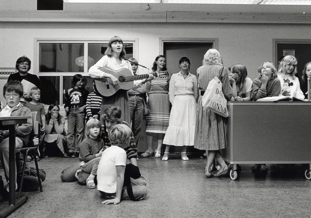 Nya Kårstaskolan vid Bergsjövägen första skoldagen höstterminen 1982 .  Skolbarnen med föräldrar och skolans personal har samlats i matsalen. En av lärarna sjunger och spelar gitarr