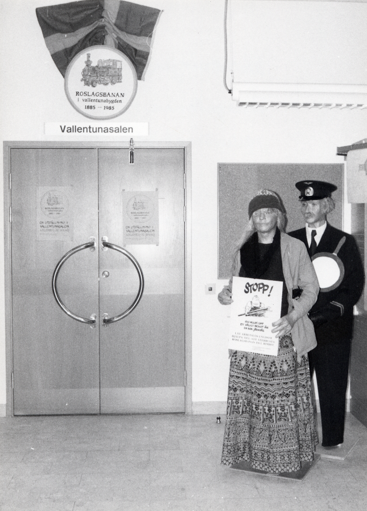 Utställningen Roslagsbanan i vallentunabygden 1885-1985 visades i Vallentunasalen 26 november 1985-7 januari 1986. Utställningen visade banans utveckling under 100 år, från invigningen 18 december 1885 till våra dagar. Hembygdsföreningarna längs banan arbetade flera år före jubiléet med att samla material i Dokumentationsgruppen för Roslagsbanan och bygden. Materialet sammanställdes av Eric Jarneberg och gavs ut i bokform Roslagsbanan och bygden i september 1985. Utställningen i Vallentunasalen sammanställdes av Eva Rahmqvist, Gunhild Hillbo och Gun Hillbo. Dockorna har Eva Rahmqvist tillverkat, kläder och material har medlemmar i vallentunasektionen av dokumentationsgruppen bidragit med. Föreningen Rädda Roslagsbanan har bidragit med informationsmaterial från många års kamp för banans bevarande samt deltog även med en egen vägg i utställningen. Bilden visar ingången till Vallentunasalen från foajén med en stins och en demonstrerande flicka med plakat med text 'Riv hellre upp ett dåligt beslut än en bra järnväg'