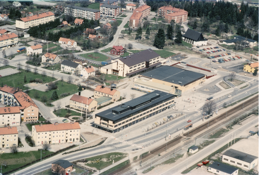 Flygfoto över Vallentuna centrum från mitten av 1980-talet. I bildens mitt Tunahuset (invigs 1973) med svart tak, bakom syns Vallentuna kommunalhus med flyglar och mellandel som rivs 1986. Bakom kommunalhuset ligger kommunalhusparken. I förgrunden till vänster om tågspåret går Banvägen diagonalt. Den röda villan bakom förvaltningshuset Tärningen (byggt 1982) uppe till höger om kommunalhuset är en av de två syskonvillorna, den norra, som rivs 1994. Den södra villan revs i början av 1980-talet.
