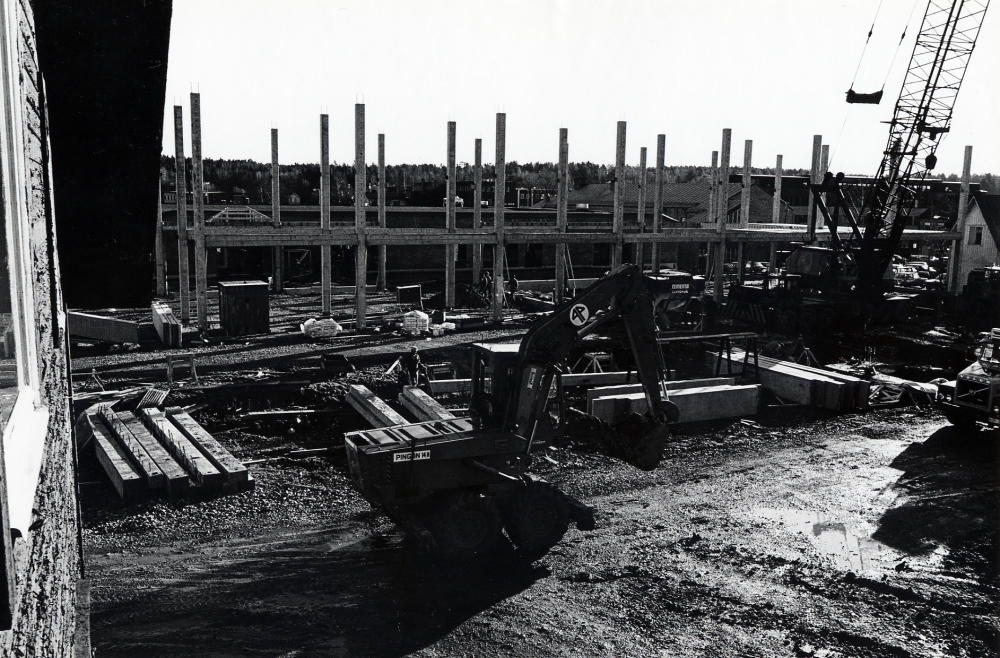 Förvaltningshuset  Tärningen  byggs.  Stommen har rests.  Bakom betongstolparna syns det nya  Domus  samt gamla  konsumhuset  och gaveln på affären  Kära Barn  I förgrunden står en  grävskopa