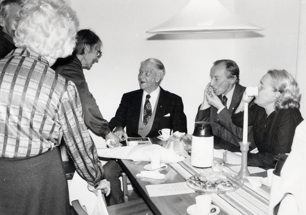 Josef Anderssons bok "Skola i brytningstid" presenteras i Össeby-Garns församlingshem. Vid bordets kortsida sitter författaren och skakar hand med Rosa Johansson. T h om Josef Andersson sitter Ralph Uddman, Folkbildningsförbundet och kulturnämndens ordförande Lillian Glav. Med ryggen mot kameran t v står Signe Andersson