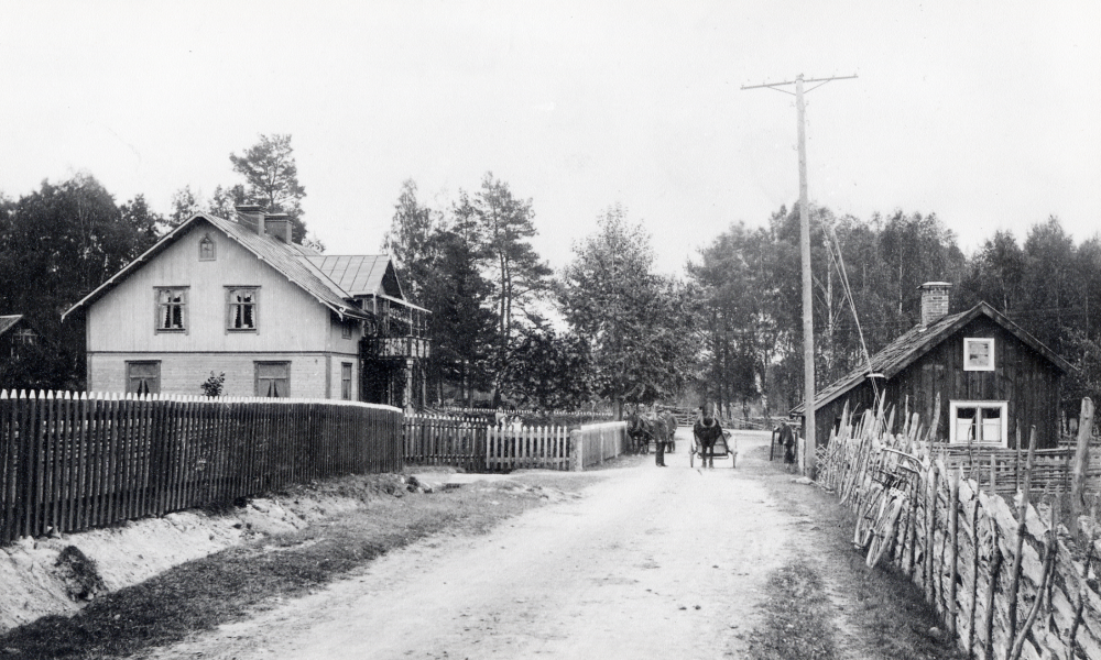Ekebydals handelsbod sommaren 1906. Handlaren Conrad Österberg byggde handelsboden på 1890-talet. På vägen i bildens mitt står två hästar med vagnar samt en cykel står lutad mot gärdesgården på vänster sida av vägen. Se: Lanthandlare i vallentunasocknarna vid Roslagsbanan
