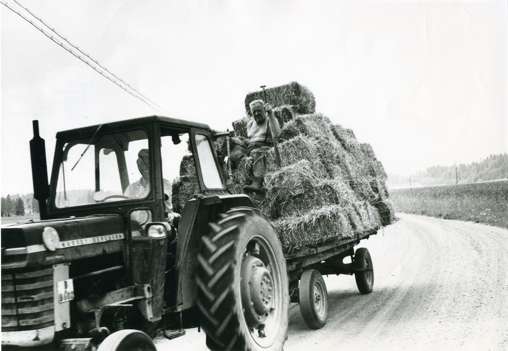 Transport av höbalar med traktor och vagn. Ingvar Österberg kör traktorn, Fritz Johansson sitter på hölasset.
•Fotografiet ingår i bildserien ”Vardagsliv i Vallentuna kommun” av Rachael Gough-Azmier & Gunilla Blomé, Haga Studios HB.