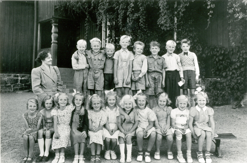 Lärarinnan Karin Norberg, maka till postmästaren i Vallentuna, tillsammans med barnen i småskolan, fotograferade utanför Röda skolan i Hjälmsta.. I bakre raden näst längst t h står Elwe Nilsson.