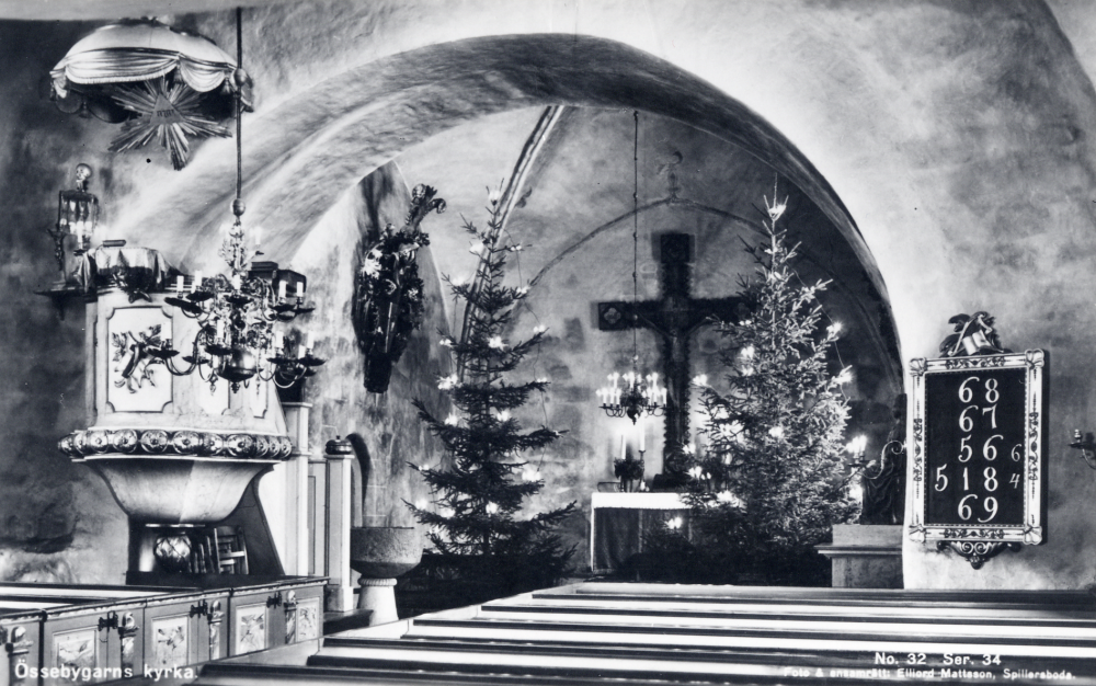 Interiör från Össeby-Garns kyrka vid jultid. I koret står två stora julgranar med elektrisk belysning. Uppgift om fotoår saknas, men troligen första delen av 1900-talet.