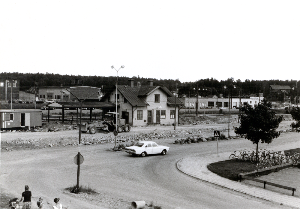 Vallentuna centrum tidigt 1970-tal, före augusti 1972. Korsningen Allévägen / Banvägen med Vallentuna station och stationshuset mitt i bild. Vägarbete / schaktning med traktor pågår framför stationshuset. En arbetsbod är uppställd till vänster i bild. Stationshuset rivs sedan, i augusti 1972. Till höger syns ett hörn av parken framför det gamla kommunalhuset. En bil kör söderut längs Banvägen.