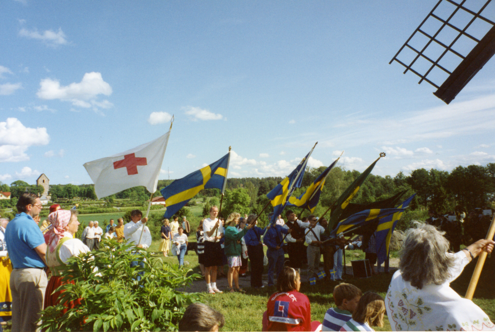 Sveriges nationaldag och svenska flaggans dag firas den 6 juni 1993. Deltagarna har marscherat från Kommunalhusparken till Väsby kvarn med Svenska flaggan och Röda korsets flagga. Kvarnens ena vinge syns till höger i bild. Längst bort till vänster skymtar Vallentuna kyrka.
Foto och uppgifter: Fredrik Andersson.
