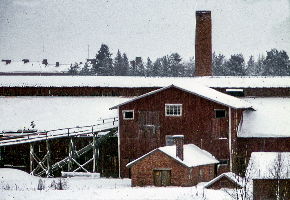 Snötäckta tak på Åby tegelbruk sett från öster. Åby tegelbruk byggdes 1927 av fabrikör C J Gustavsson. Driften lades ner 1968 och bruket brann ner i september 1970. Marken såldes till Riksbyggen.
Bildserie: Vallentuna på 1960-talet.