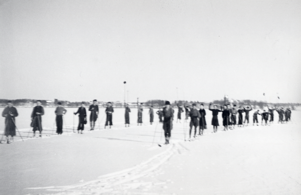 Skolbarn från Hjälmsta skola på skidutflykt, fotograferade på Vallentunasjön på väg till Kragsta för att öva backåkning.  I bakgrunden Vallentuna kyrka