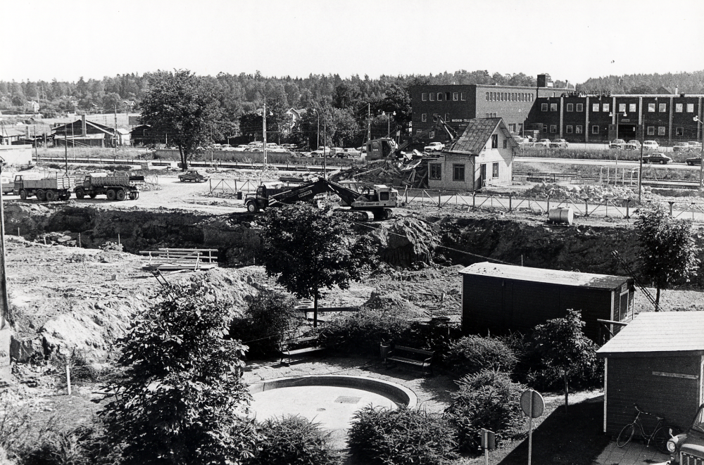 Anläggningsarbeten har påbörjats inför byggstarten av det nya förvaltningehuset Tunahuset.   I bakgrunden rivs Vallentuna järnvägsstation.  I förgrunden t v skymtar gaveln på kommunalhuset.  Bakom stationshuset är Maskin-Tellus fabrik.  I förgrunden syns en liten damm med plantering